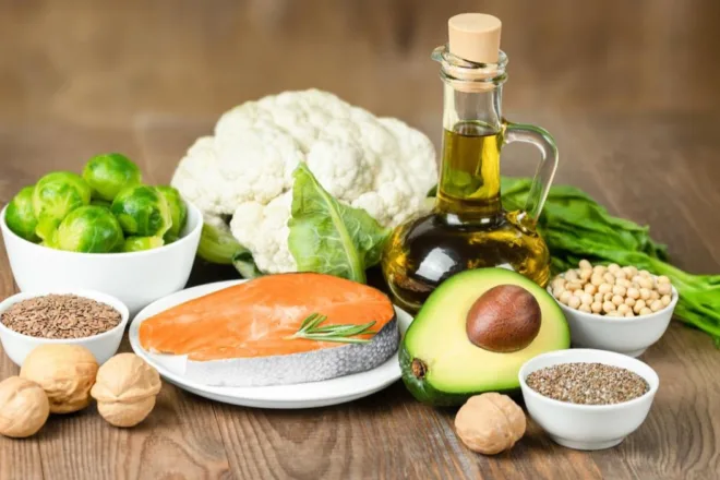 Assorted low-carb foods including salmon, avocado, olive oil, cauliflower, Brussels sprouts, nuts, and seeds on a wooden table.