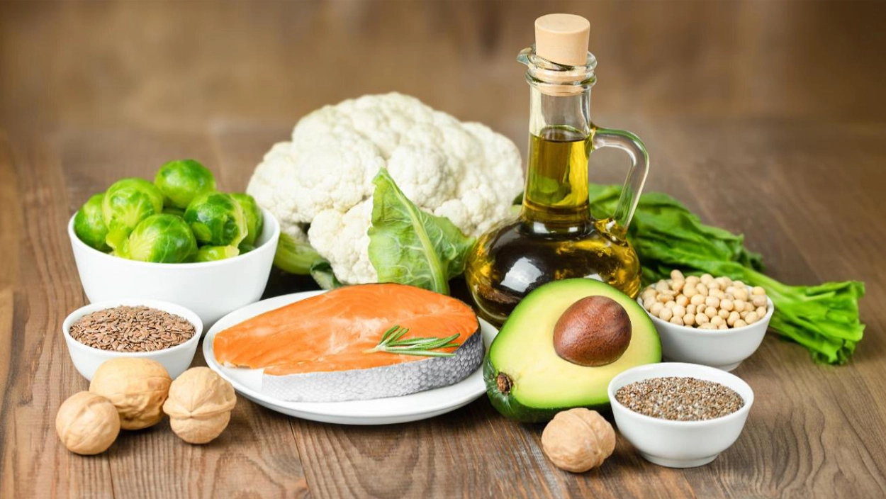 Assorted low-carb foods including salmon, avocado, olive oil, cauliflower, Brussels sprouts, nuts, and seeds on a wooden table.