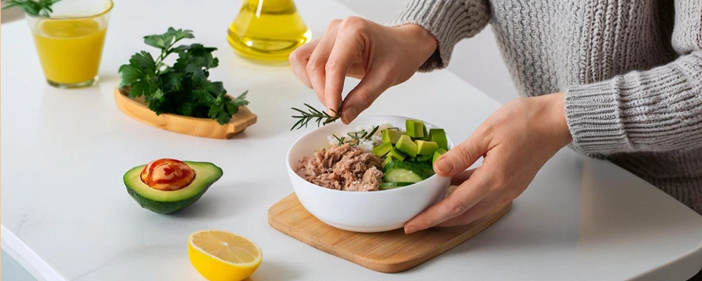 Person preparing a low-carb meal with tuna, avocado, cucumber, and fresh herbs on a kitchen counter.