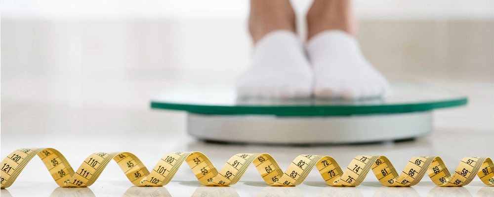 Person checking weight on a digital scale with measuring tape on the floor, using it later for a quick BMI calculator.