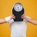 Man holding a weight scale in front of his face on yellow background