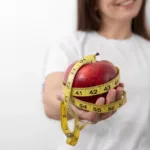 Woman holding a red apple wrapped in a yellow measuring tape, illustrating apple-shaped body and BMI analysis