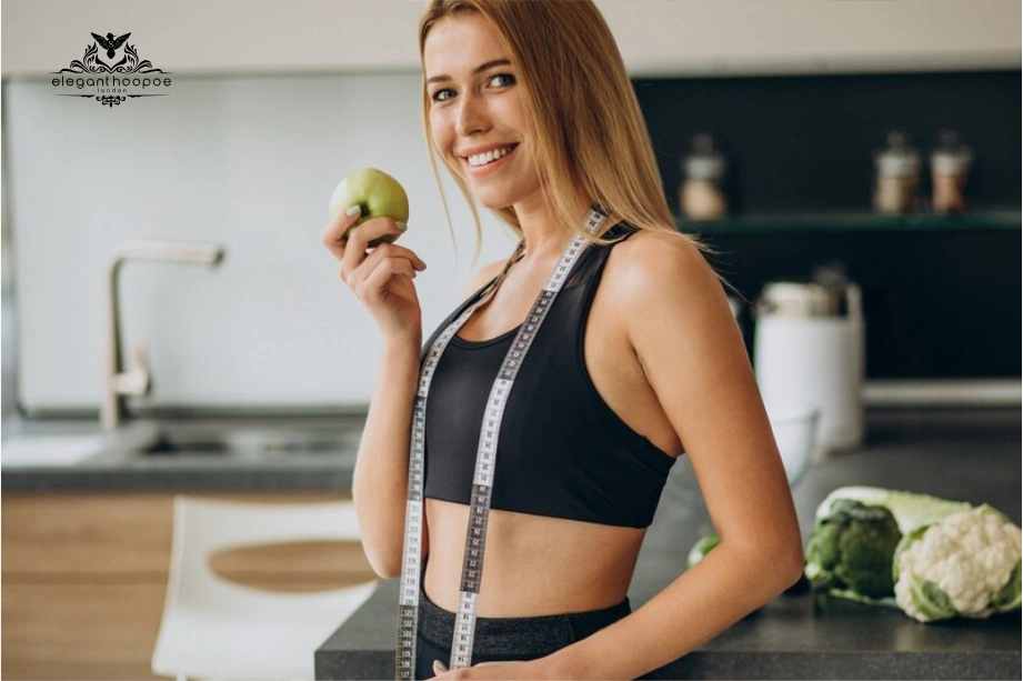 Fit woman holding an apple and wearing a measuring tape around her neck in a kitchen.