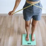 Woman on scale crossing tape around waist, DIY belly-fat test