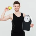 Smiling man holding weighing scale and green apple to support body mass index awareness.