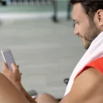 Male bodybuilder checking BMI app after workout, towel on shoulder.
