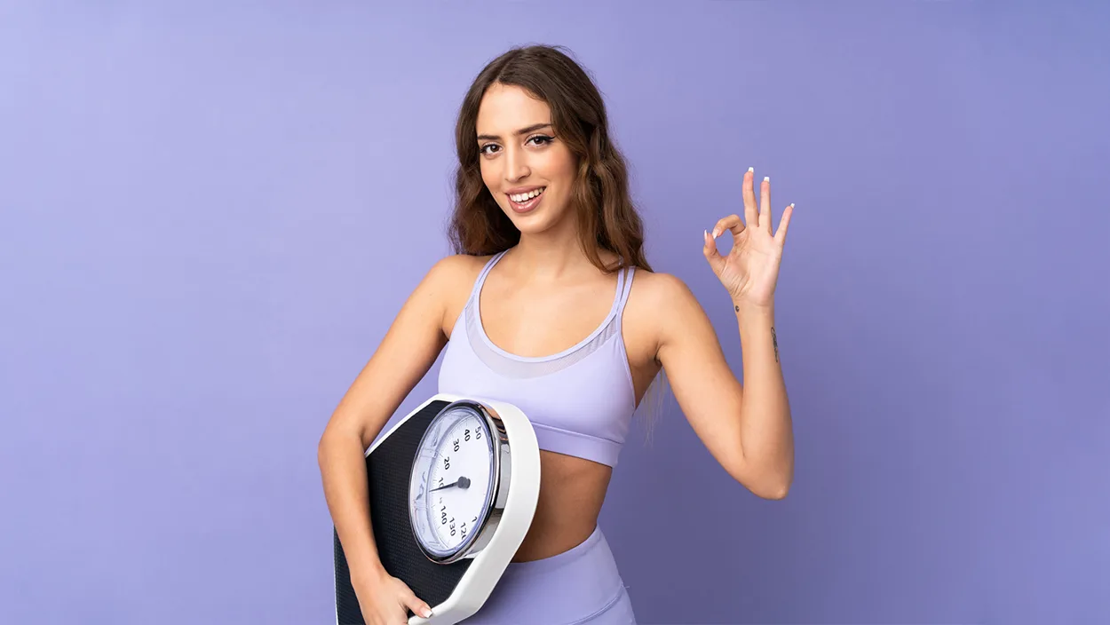 Woman in purple athletic wear holding a scale and making an OK gesture, symbolizing success with the ideal weight calculator for men and women