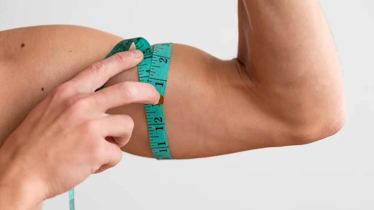 Close-up of a person measuring their flexed bicep with a green tape measure to assess muscle mass for fitness tracking