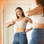 Woman in white tank top and jeans giving a thumbs-up in front of a mirror, evaluating her body frame size