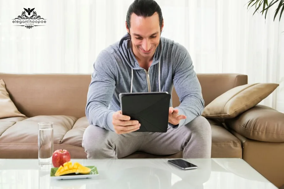 Man sitting on couch using a tablet to check BMI online, with glass of water, apple, and mango slices on a table.