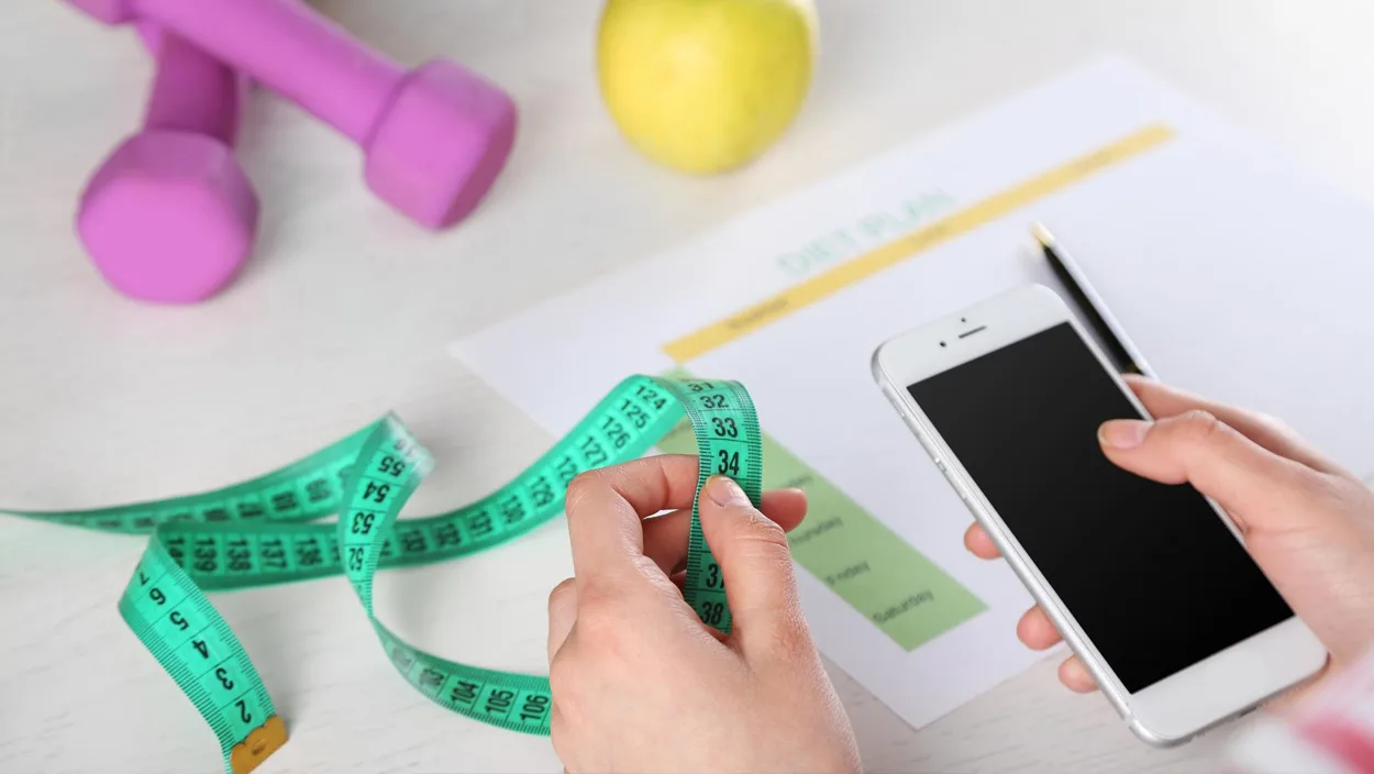 Person holding a smartphone for biomass index calculator and tape measure beside a diet plan, apple, and dumbbells