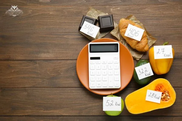 Calculator on an orange plate surrounded by foods labeled with calories: chocolate (546 kcal), croissant (406 kcal), red pepper (27 kcal), pear (32.2 kcal), squash (28 kcal), apple (52 kcal)
