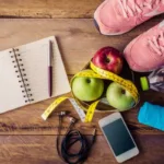 Notebook, pen, apples, measuring tape, pink sneakers, water bottle, smartphone, earphones, and towel on wooden surface