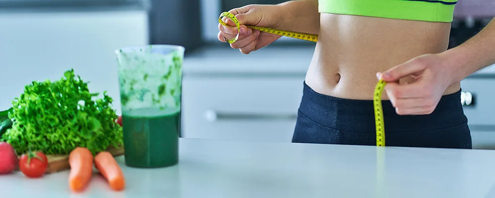 A person measuring waist circumference with a tape measure, surrounded by fresh vegetables and a green smoothie, symbolizing health tracking and achieving biomass index goals