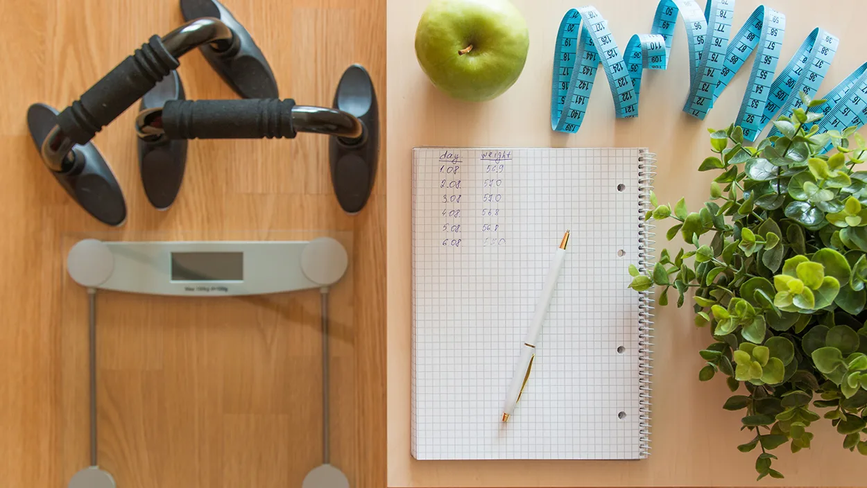 A flat lay of health tracking tools including a digital scale, push-up bars, a green apple, a blue measuring tape, a notebook with weight-tracking entries, a pen, and a potted plant.
